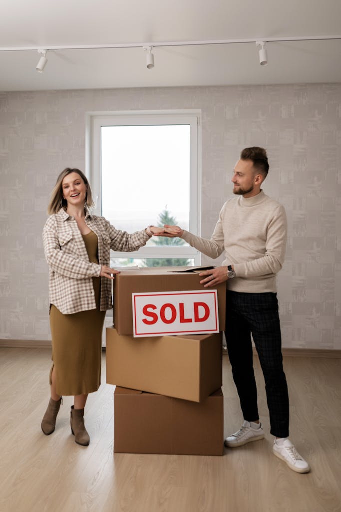 Smiling couple celebrating their new home purchase with sold sign and cardboard boxes.